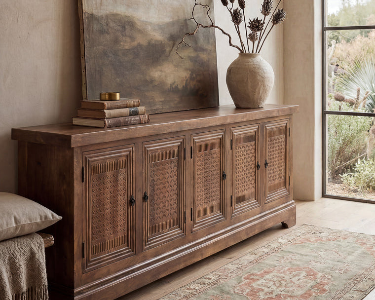 A rustic wooden sideboard cabinet with woven cane doors, styled in a warm, neutral-toned room with a large landscape painting, a ceramic vase with dried branches, and a patterned rug.